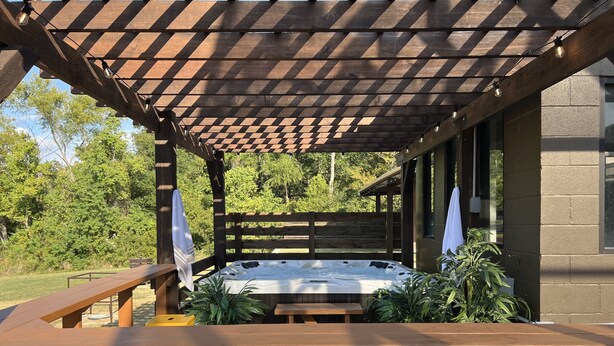 Een terras buiten met een houten pergola, een jacuzzi, gele plastic stoelen en potplanten.