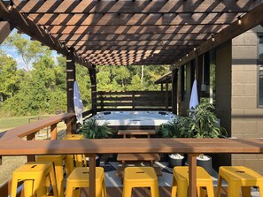 An outdoor patio with a wooden pergola, a hot tub, yellow plastic stools, and potted plants.