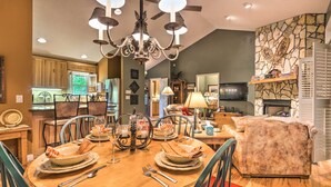 A dining area with a round table set for four, surrounded by chairs. A chandelier hangs from the ceiling, and there is a fireplace in the background.