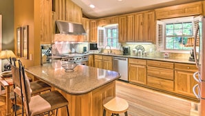 A kitchen with wooden cabinets, granite countertops, a stainless steel range hood, a built-in oven, a dishwasher, and a window with a view of greenery.