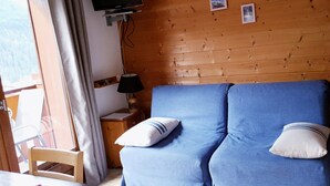 Une pièce avec un canapé bleu, deux coussins, une petite table avec une lampe et un téléviseur fixé au mur.