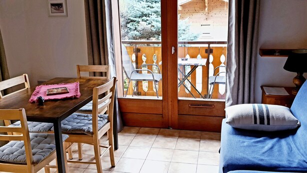 Une pièce chaleureuse avec une table à manger en bois, des chaises, un canapé et une vue sur un balcon aménagé avec du mobilier d’extérieur.