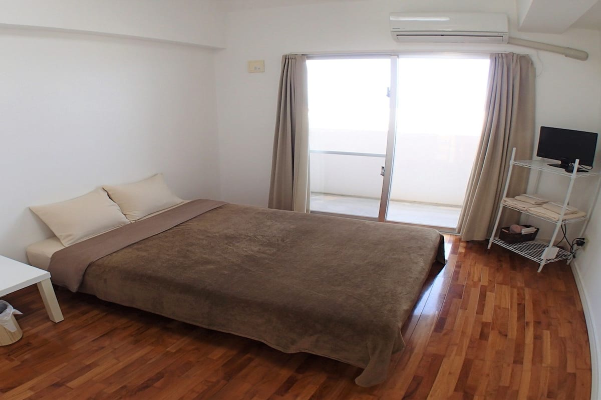 A bedroom with a bed, a desk, a chair, a TV on a stand, and a window with curtains.