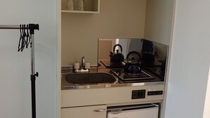 A compact kitchen with a sink, stove, and a small refrigerator. There are shelves above the sink holding cups and glasses. - Lagoon Palace (Kadena)