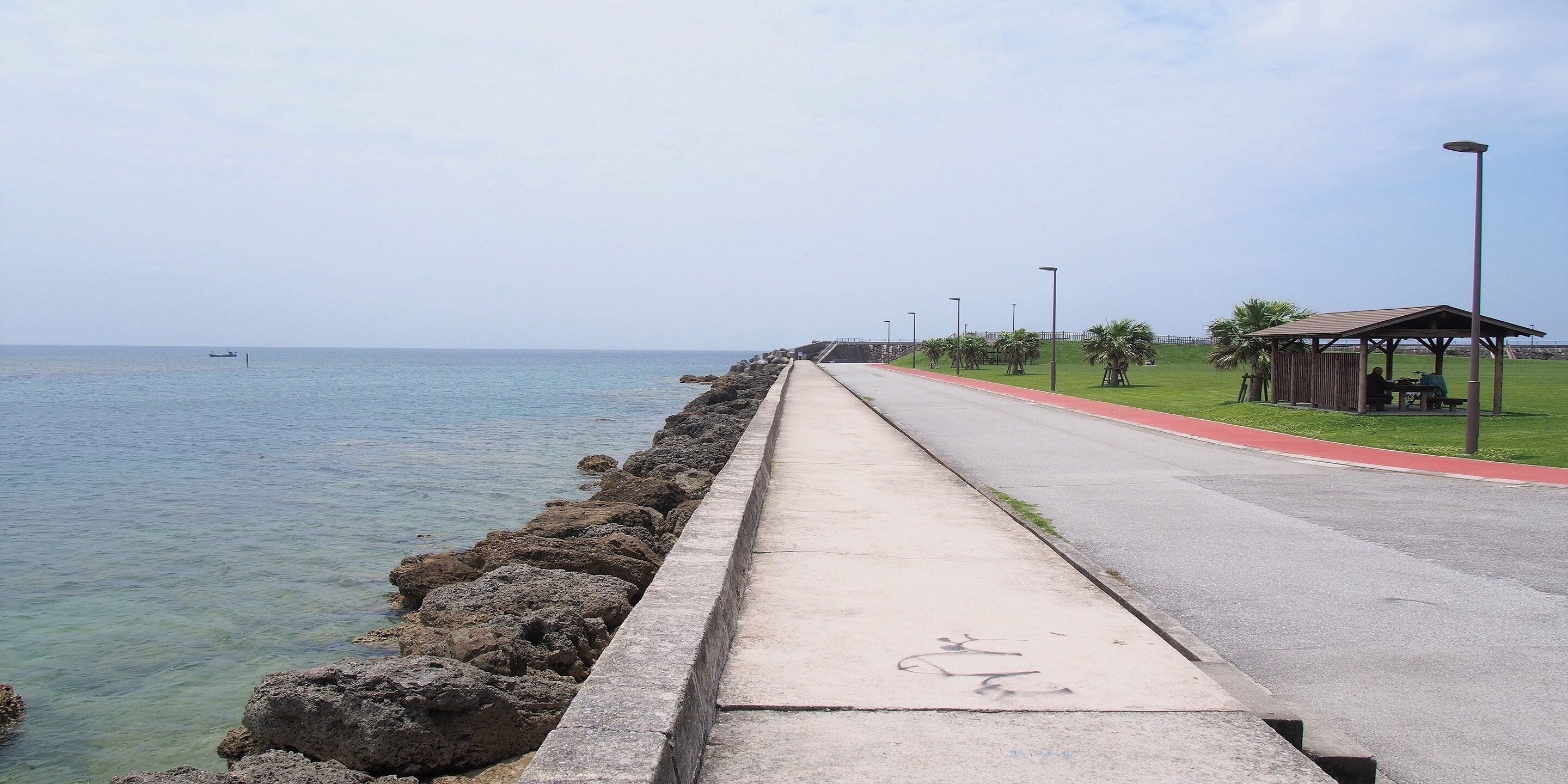 Un paseo marítimo con sendero de hormigón, carril bici rojo y un pequeño refugio con bancos.