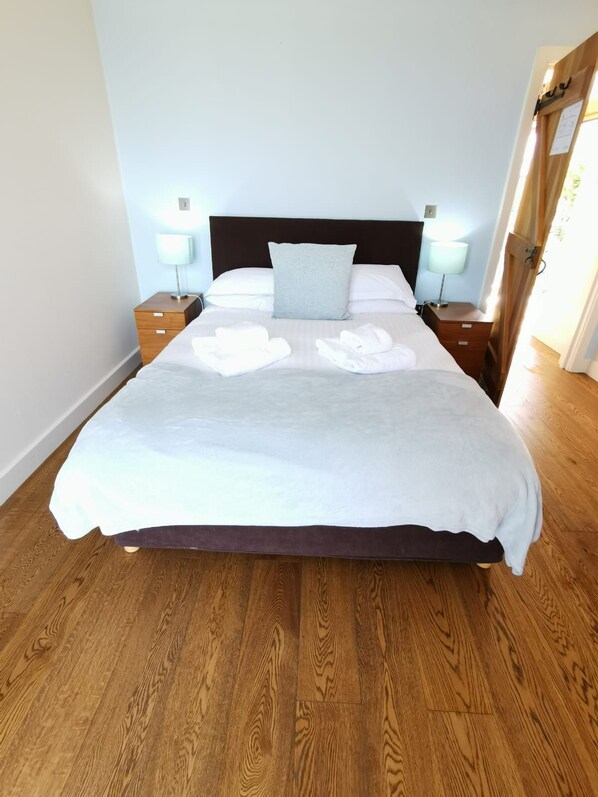 Una cama bien tendida con sábanas blancas y almohada gris, mesitas de noche de madera con lámparas, y suelo de madera.