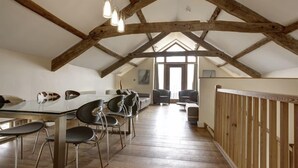 Un loft amplio con vigas de madera a la vista, una zona de comedor con mesa de cristal y sillas de metal, y vistas al exterior a través de grandes ventanales.