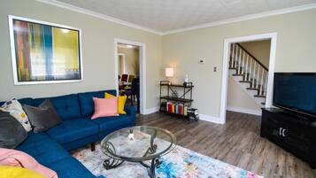 A living room with a blue sofa, a glass coffee table, a colorful rug, a TV, and a bookshelf.