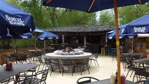 Un espace extérieur aménagé en terrasse, avec des tables et des chaises rondes, protégé par des parasols bleus arborant la marque « SAMUEL ADAMS », et un foyer central en pierre.