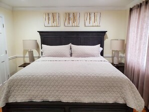 A neatly made bed with a quilted white bedspread, flanked by two bedside tables with lamps, in a room with cream walls and a dark wood headboard. - 1118-1 First roll out loud (Queens)