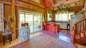 A rustic wooden interior with a kitchen area, a dining table with red chairs, and a large window providing natural light. - Treehouse Vibe Near Hilo & Volcano w/ AC, BBQ, W&D & Ocean Cliffs - Keaau (Keaau)