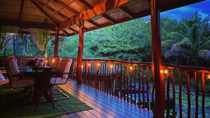A wooden deck with a railing, chairs, and a table, overlooking a lush green landscape. - Treehouse Vibe Near Hilo & Volcano w/ AC, BBQ, W&D & Ocean Cliffs - Keaau (Keaau)
