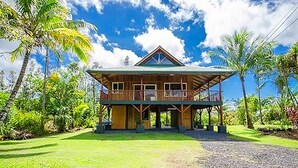 A two-story wooden house with a balcony, surrounded by lush greenery and palm trees. - Treehouse Vibe Near Hilo & Volcano w/ AC, BBQ, W&D & Ocean Cliffs - Keaau (Keaau)