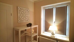 Ett rum med ett element, ett vitt konsolbord med en växt, en dekorativ väggdekoration och ett fönster med persienner.