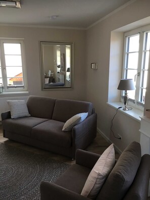 En stue med sofa, en mindre lænestol, et stort spejl, en lampe og et vindue med persienner.