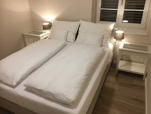 A neatly made twin bed with white linens, two bedside tables with lamps, and a window with blinds.