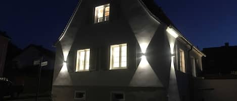 A dark-colored house with two visible windows, one on the upper floor and one on the ground floor, illuminated by exterior lighting.