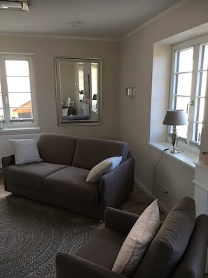 Ein Wohnzimmer mit einer Couch, einem Sessel, einem großen Spiegel, einer Lampe und einem Fenster mit Jalousien.