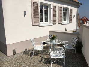 A patio with a round table and four chairs, a potted plant, and a building with white walls and shutters.