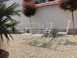 Ein weißes Metall-Garnitur mit Tisch und Stühlen auf Kies, mit zwei kleinen Bäumen und einem Blumentopf in der Nähe.