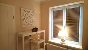 Ein Zimmer mit Heizkörper, einem weißen Beistelltisch mit Pflanze, einer Wanddekoration und einem Fenster mit Jalousien.