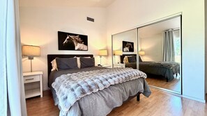 Un dormitorio con un espejo grande, una cama con una manta gris, dos mesitas de noche con lámparas y un cuadro de un caballo colgado en la pared.