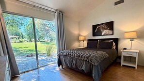 A bedroom with a large sliding glass door leading to a garden, a bed with a grey duvet, two bedside lamps, and a framed picture on the wall.