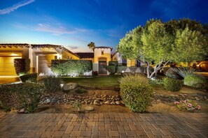 A well-lit residential property with a paved driveway, landscaped garden, and a two-story house with a tiled roof. - Bliss: Pool, Spa, Pool Table, Ping Pong, Fire Pit! (Indio)