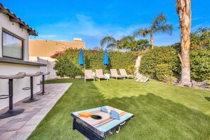 A backyard with a lawn, lounge chairs, a table with a board game, and a tall palm tree. - Bliss: Pool, Spa, Pool Table, Ping Pong, Fire Pit! (Indio)