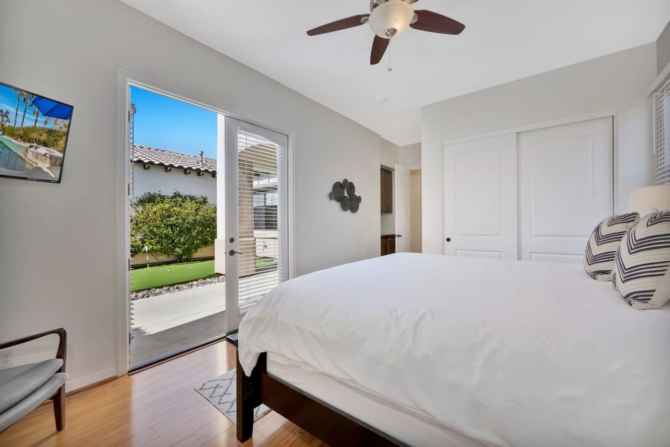 A bedroom with a bed, a ceiling fan, a door leading to a balcony, and a view of a building with a tiled roof.