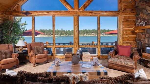 A log cabin interior with a large window overlooking a lake, furnished with leather chairs and a wooden table. - Lakefront Luxury Private Dock, Spa & Game Room (Big Bear Lake)