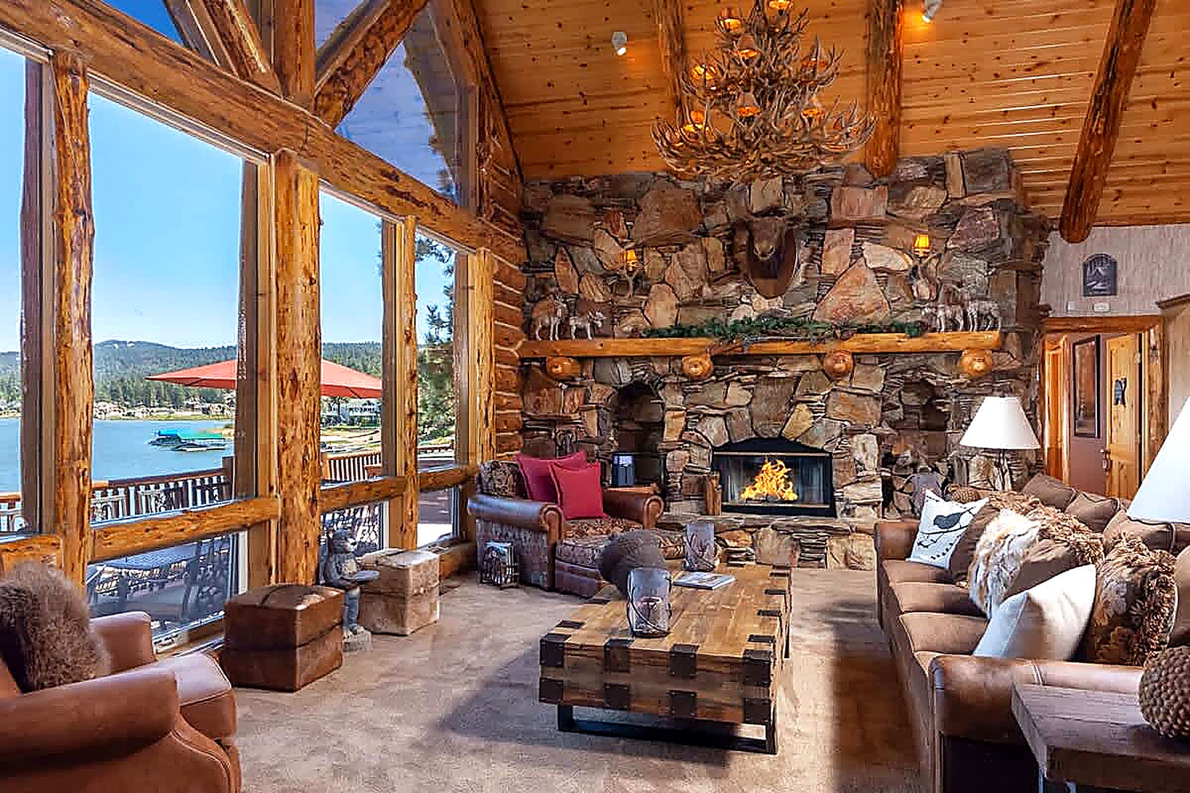 Interior de una cabaña de troncos con chimenea de piedra, amplias ventanas con vistas a un lago y muebles rústicos.