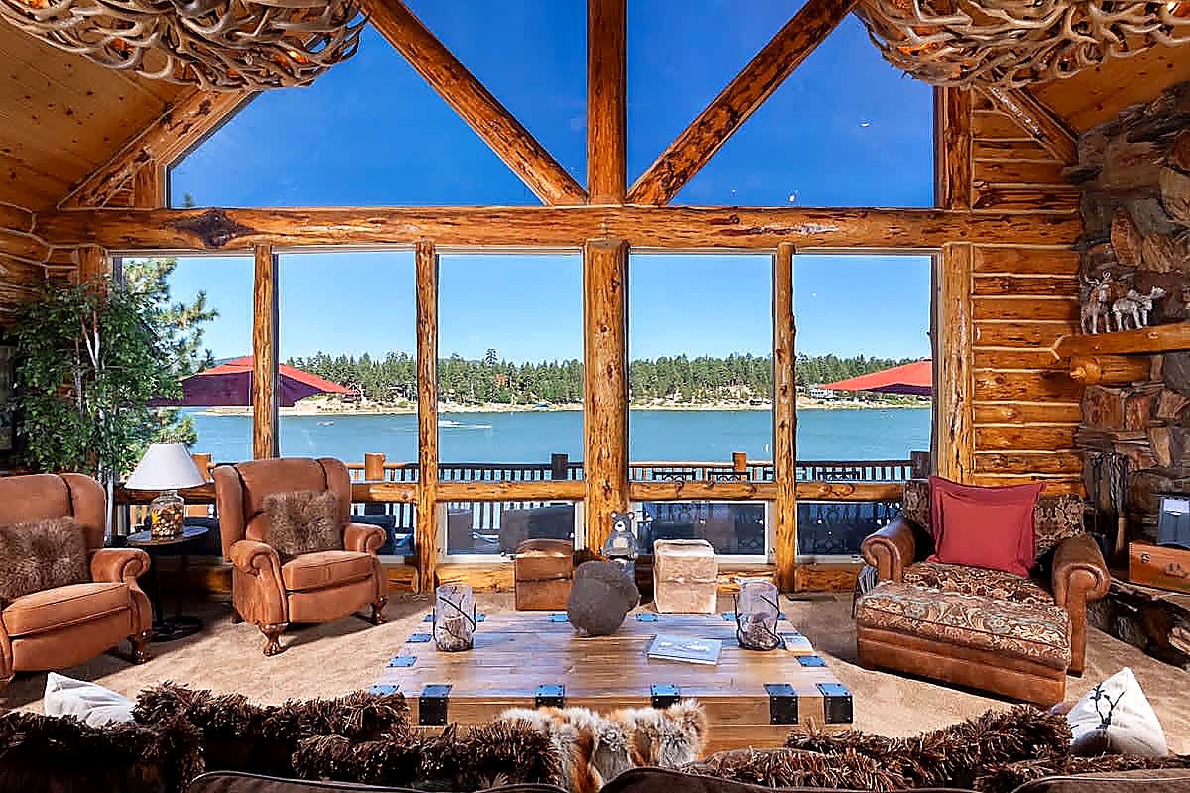 Interior de una cabaña de madera con un ventanal que da a un lago y un bosque.