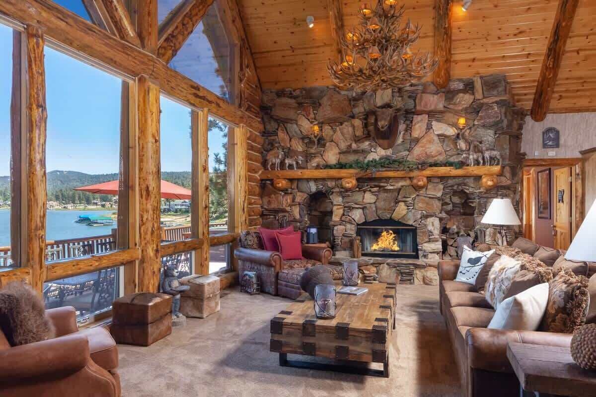 Interior acogedor de una cabaña con chimenea de piedra, amplias ventanas con vistas a un lago y vigas de madera rústicas.