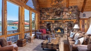 A cozy lodge interior with a stone fireplace, large windows overlooking a lake, and rustic wooden beams. - Lakefront Luxury Private Dock, Spa & Game Room (Big Bear Lake)