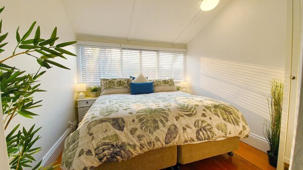 Un dormitorio con cama, ventana con persianas, mesita de noche con lámpara y una planta en maceta.