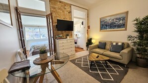 Ein Wohnzimmer mit einer Couch, einem Glastisch, einem an der Wand befestigten Fernseher, einem Gemälde und einer Zimmerpflanze.