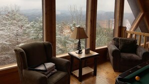 A cozy room with large windows offering a view of snow-covered trees and mountains. There is a small table with a lamp and a chair with cushions. - MTN VIEWS! True Mtn Wood Cabin-Pool Tble-6x parking-main level master-wifi (Sevierville)