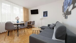 A modern living room with a grey sofa, a dining table with chairs, a small round table, a flat-screen TV, and artwork on the wall.