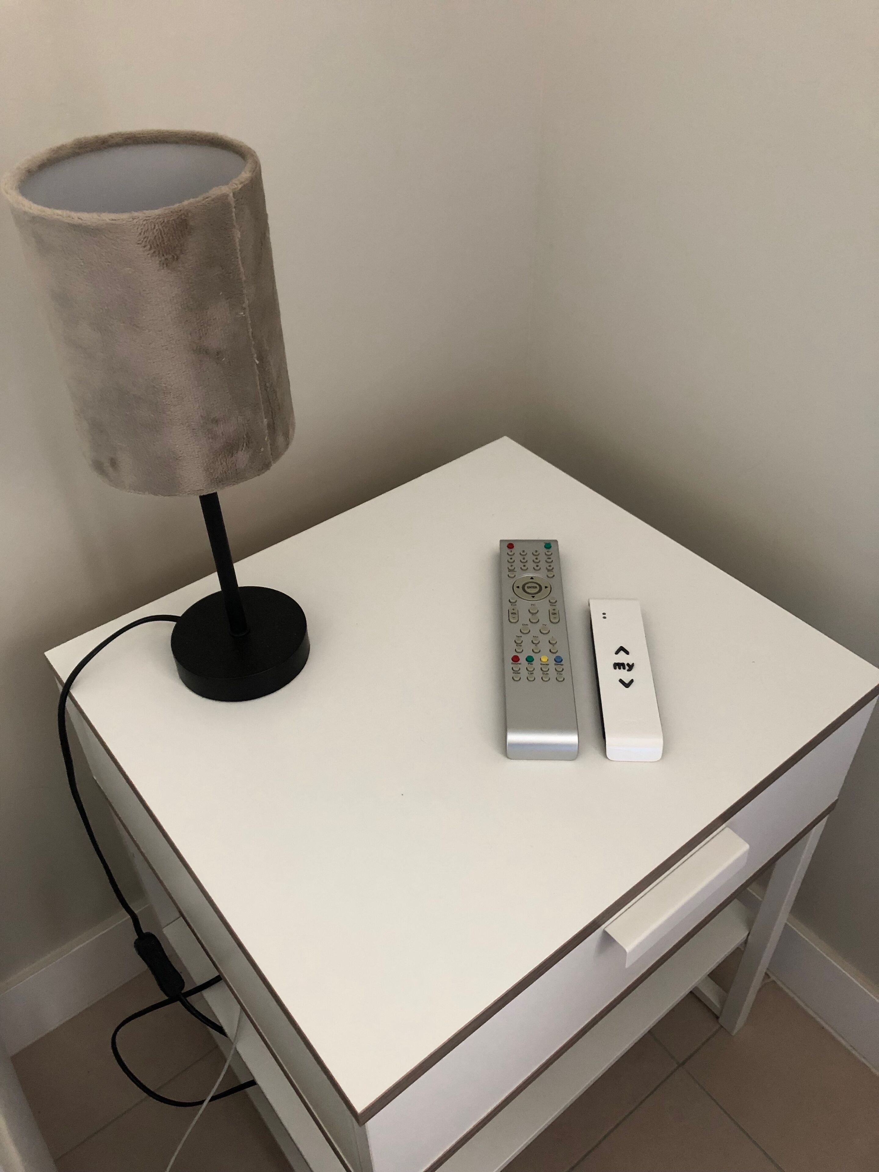 A small white bedside table with a lamp and two remote controls.