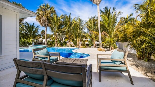 A pool area with lounge chairs and a small table, surrounded by palm trees and a clear blue sky. - At Last: Ground-level, pool, hot tub, canalfront, dock (Anna Maria)