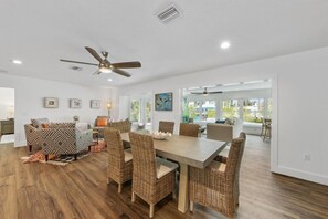 A spacious dining area with a large table and wicker chairs, a ceiling fan, and a view of the outdoors through large windows.