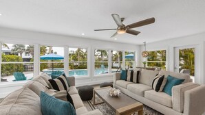 A spacious living room with a beige sofa set, a coffee table, and a ceiling fan. Large windows offer a view of a pool and outdoor furniture. - At Last: Ground-level, pool, hot tub, canalfront, dock (Anna Maria)