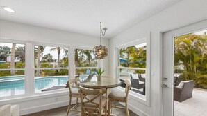 A dining area with a round table and chairs, large windows with a view of a swimming pool and greenery, and a hanging light fixture. - At Last: Ground-level, pool, hot tub, canalfront, dock (Anna Maria)