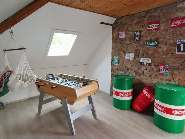 A room with a foosball table, a hanging macrame chair, and a brick wall adorned with various brand signs. - Large gite with 9 places (Lièpvre)