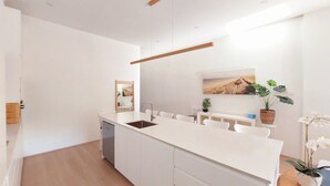 Una cocina moderna con armarios blancos, una isla central con lavaplatos y un área de comedor con sillas blancas. Hay plantas y un cuadro en la pared.