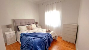Una cama bien hecha con una manta azul, una mesita de noche con una lámpara y otra mesita con una lámpara.
