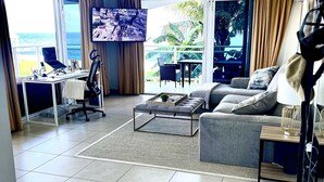 Ein geräumiges Wohnzimmer mit großem Fernseher, einer Couch, einem Couchtisch und einem Schreibtisch mit Stuhl. Große Fenster bieten Blick auf den Ozean.