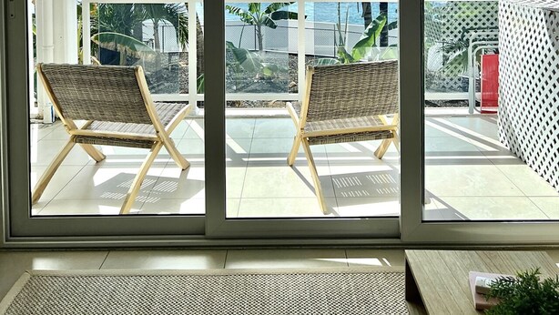 Ein Balkon mit zwei Korbstühlen und einem kleinen Tisch, mit Blick auf die Küste, Palmen und einen klaren Himmel.