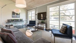 Una sala de estar moderna con un sofá, sillones, una mesa de centro y un televisor. Hay ventanas amplias y un ventilador de techo.